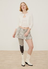 Jeune femme portant un cardigan court blanc, une mini-jupe à fleurs, des bottes blanches et une jambe gauche prothétique avec une housse noire.