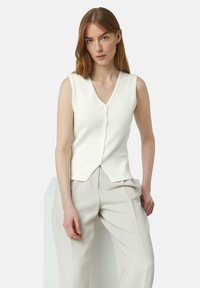 Witte mouwloze gebreide vest met V-hals, voorzien van een knoopsluiting aan de voorkant en een licht uitlopende zoom, gecombineerd met lichtgekleurde broeken.
