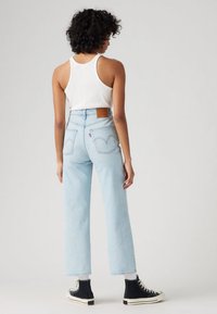 Γυναίκα με σγουρά μαλλιά φορούν λευκή αμάνικη μπλούζα, ανοιχτό μπλε τζιν ψηλόμεσο, λευκές κάλτσες και μαύρα αθλητικά παπούτσια ψηλού τύπου, στέκεται με την πλάτη.