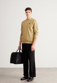 Kaki mikina s okrúhlym výstrihom a logom, kombinovaná s čiernymi širokými nohavicami. Modelka drží čiernu cestovnú tašku a stojí na svetlej podlahe.