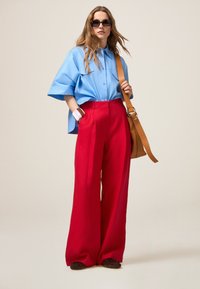 Lichtblauw oversized shirt met korte mouwen; wijde, hooggeplaatste rode broek; bruine schoudertas; gelaagde armbanden; zonnebril.