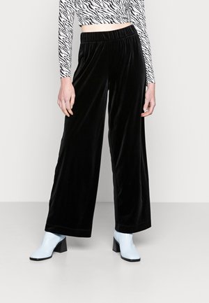 Persoon die zwarte wijde fluwelen broek draagt, een crop top met zebraprint en lange mouwen, en lichtblauwe enkellaarsjes, staand op een effen vloer.