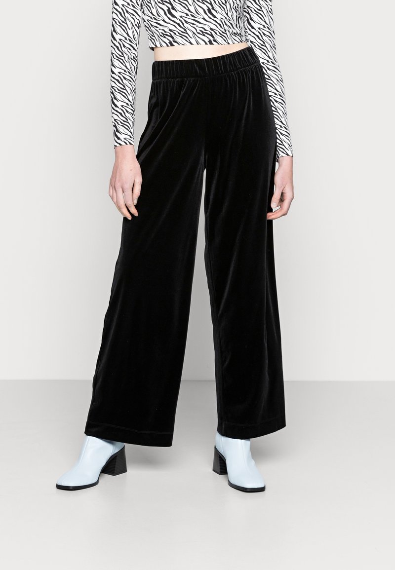 Personne portant un pantalon en velours noir à jambes larges, un crop top à manches longues imprimé zèbre et des bottines bleu clair, debout sur un sol uni.