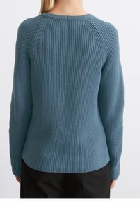 Femme portant un pull en tricot bleu vue de dos, avec des manches longues et un pantalon noir sur un fond clair uni.