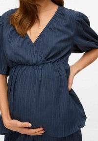 Haut bleu marine texturé avec un décolleté en V et des manches courtes bouffantes, présentant des détails froncés à la taille, porté sur un ventre arrondi.