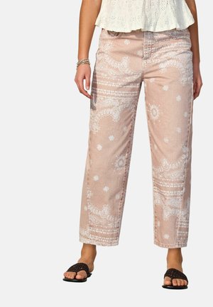 Vrouw die beige rechte broek met patroon draagt, witte korte blouse, zwarte geweven open-toe sandalen en gouden armbanden aan de linker pols.