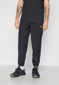 Juodos sportinės kelnės su elastinga juosmens juosta ir rankogaliais, priekyje yra subtilus Nike logotipas. Dėvimos su juodais sportiniais batais ir baltais kojinėmis.