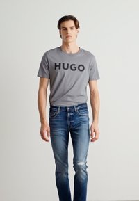Tricou din bumbac gri, cu un imprimeu îndrăzneț "HUGO" în negru, asortat cu blugi albaștri, ușor decolorați, cu un ușor efect de uzură și croi slim.
