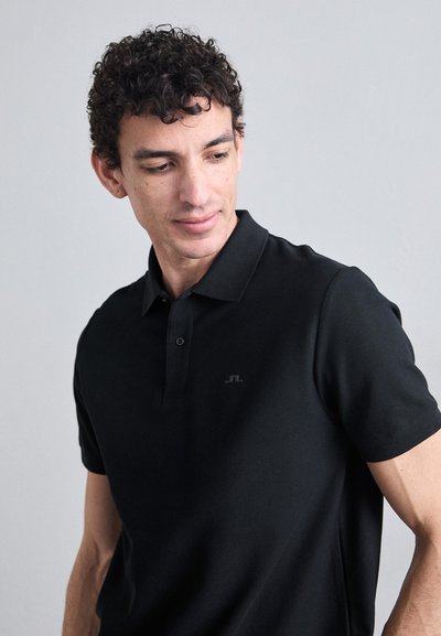 Camisa polo negra de mangas cortas, decorada con una tapeta de dos botones y un pequeño logo en el pecho. Textura suave.