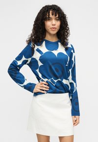 Μπλε μπλούζα με μακριά μανίκια και λουλουδάτο σχέδιο με λευκές λεπτομέρειες, κατασκευασμένη από απαλό ύφασμα. Συνδυάζεται με λευκή υφασμάτινη φούστα με υφή και εφαρμοστή γραμμή.