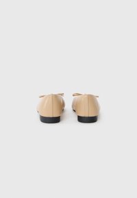 Beige ballet flats yang terbuat dari bahan sintetis halus, menampilkan ujung bulat, tumit kecil berwarna hitam, dan aksen pita di bagian belakang.