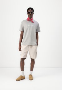 Camiseta de algodón gris con el logo "REPLAY", pantalones cortos de corduroy beige, zapatillas color marrón claro y un pañuelo rojo atado alrededor del cuello.