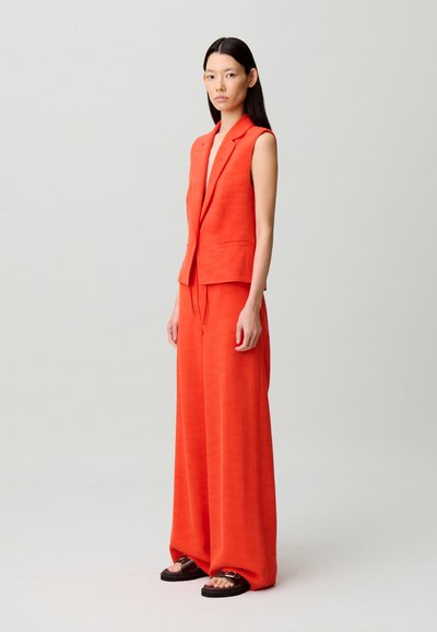 Donna in piedi che indossa un blazer senza maniche di un brillante arancione e pantaloni a gamba larga abbinati a sandali neri su sfondo semplice.