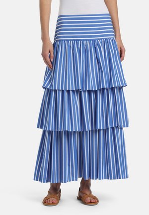 Žena v modro-bielej sukni s vertikálnymi pruhmi a hnedých sandáloch stojí pred jednoduchým pozadím.