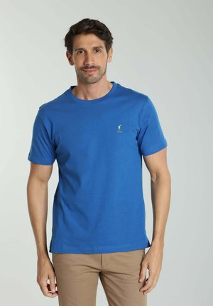 Homme aux cheveux courts et foncés portant un t-shirt bleu à manches courtes et un pantalon beige sur un fond clair uni.