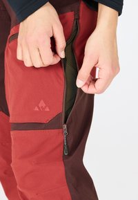 Pantalons d'extérieur rouges et marron foncé avec poches zippées. Caractéristiques d'un tissu texturé et d'une doublure en maille visible à l'intérieur de la poche.