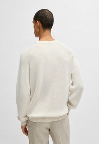 Heller cremefarbener, gerippter Pullover mit rundem Ausschnitt und Raglanärmeln. Weiche Textur, lockerer Schnitt, kombiniert mit hellgrauen Hosen.