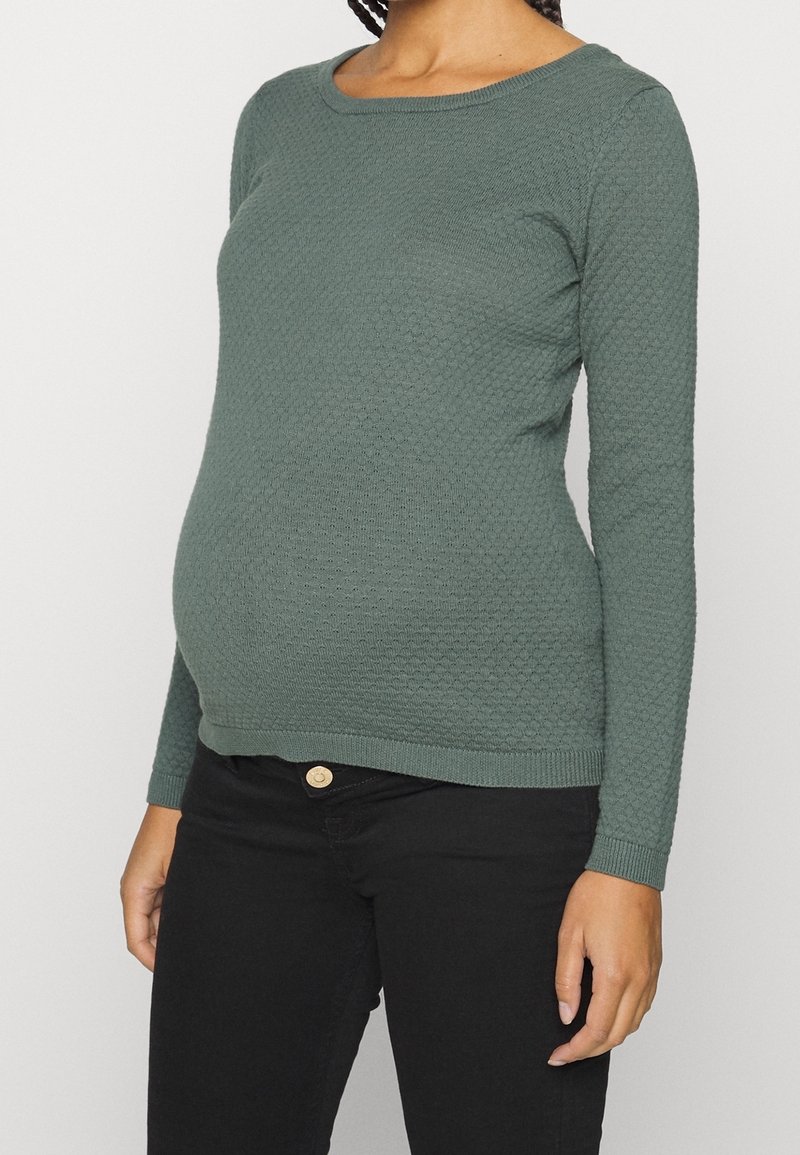 Personne enceinte portant un haut vert texturé à manches longues et un pantalon noir, visible des épaules aux hanches sur un fond uni.