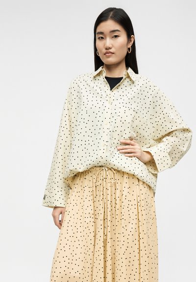 Γυναίκα με μακριά μαύρα μαλλιά που φοράει χρυσούς κρίκους στα αυτιά, μια κρέμ πουκάμισο με πουά και φούστα με κορδόνι και πουά που ταιριάζουν.