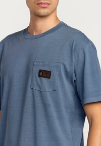 Blå bomulds t-shirt med rund halsudskæring og korte ærmer. Har en brystlomme med en mørk label og syningsdetaljer. En smule struktureret stof.