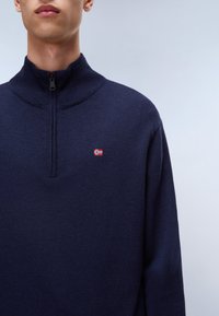 Marinefarbener Pullover mit hohem Kragen, halbseitigem Reißverschluss, gerippten Bündchen und einem kleinen gestickten Flaggenmotiv auf der Brust. Weiche Stricktextur.