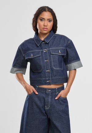 Naine punutud juustega, kes kannab lühikese varrukaga tumedat teksajakki ja sobivaid laiade säärega teksapükse, käed taskutes.