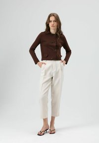 Bruine, nauwsluitende jas met lange mouwen, knoopsluiting en twee voorzakken, gecombineerd met crèmekleurige, geplooide, enkellange broek en zwarte sandalen.