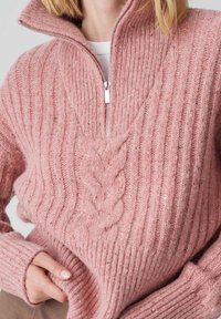 Femme portant un pull en maille côtelée rose avec un motif central en torsade et un col demi-zip, tenant l'ourlet du pull d'une main.
