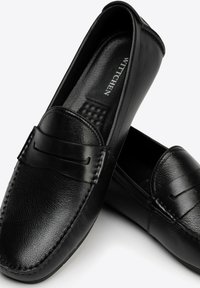 Mocasines de cuero negro con una textura suave, que presentan un diseño cosido y una banda decorativa en la parte delantera. Forma ergonómica y flexible.