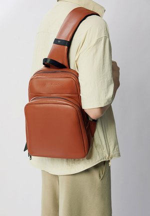 Brown leather backpack with two zippered compartments, adjustable strap, and embossed brand logo. Smooth texture and structured shape.