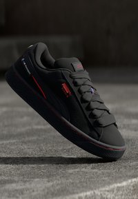 Black Puma Suede XL sneaker with BMW M Motorsport logo and red accents, suspended slightly above a concrete surface in soft light.