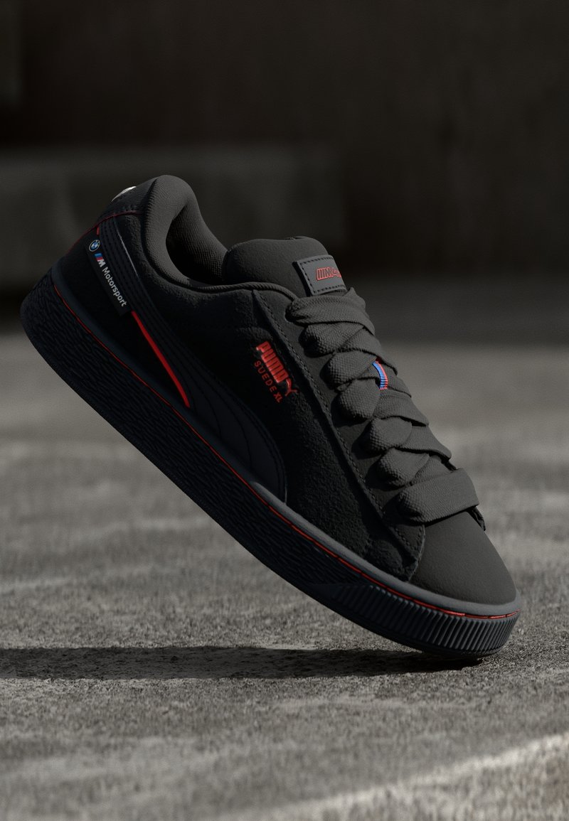 Black Puma Suede XL sneaker with BMW M Motorsport logo and red accents, suspended slightly above a concrete surface in soft light.