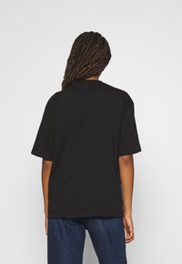 Person med flätat hår som bär en enkel svart t-shirt med kort ärm och blå jeans, sedd bakifrån mot en vit bakgrund.