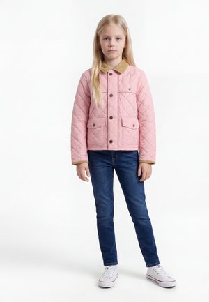 Joven chica de pie, vistiendo una chaqueta acolchada rosa con cuello marrón claro, vaqueros azules y zapatillas blancas sobre fondo blanco.