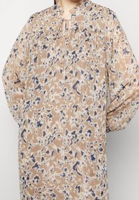 Brun blommig mönstrad blus med krage, långa ärmar och knytdetalj vid halsen. Tillverkad av ett lätt och strukturerat tyg.