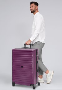 Maleta rígida morada con diseño estriado, dos ruedas de rodadura suave y un mango extensible, siendo llevada por una persona con ropa ligera.