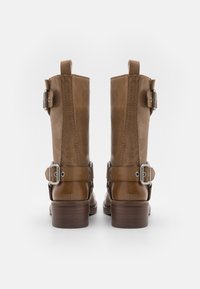 Par av bruna läderstövlar som når till vaden, med spännen vid anklarna och toppen, staplade träklackar, sedd från baksidan.