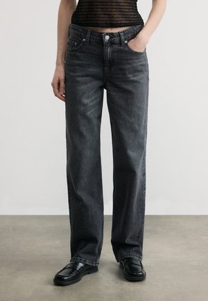 Personne portant un jean noir droit, des mocassins en cuir noir et un haut en maille noire avec un texte répété, debout avec une main dans la poche.