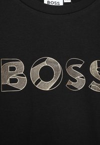 Svart T-shirt med ordet "BOSS" i stora, guld- och konturerade bokstäver med varierande linjemönster, mot en slät tygtextur.