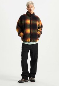 Fleecevest in zwart, oranje en groen geruit patroon, met een ritssluiting aan de voorkant en een hoge kraag, gecombineerd met zwarte broek en robuuste schoenen.