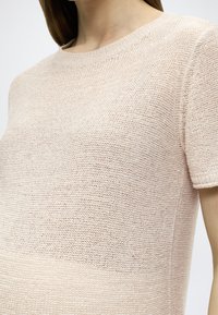T-shirt en maille rose clair, doté d'un col rond et de manches courtes. La texture est en maille ajourée avec un motif rayé subtil.