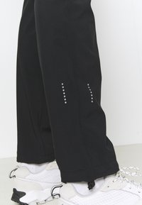 Black pants made of a smooth, stretchy fabric. Feature reflective dots on the sides and drawstrings at the hem. Paired with white sneakers.