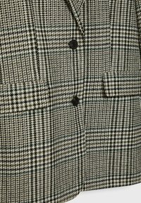 Houndstooth-kuvioitu takki mustilla napeilla ja kahdella etutaskulla, väreinä musta, valkoinen ja oliivinvihreä.