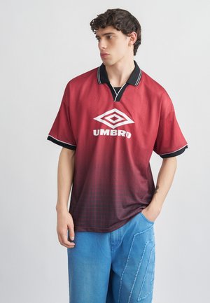 Mlad moški, ki gleda v stran, oblečen v rdečo in črno polo majico Umbro ter modre hlače z nalepko, z roko v žepu na belem ozadju.