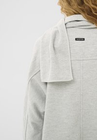 Világosszürke pulóver, köralakú nyakkivágással, amelynek vállain egy hurkolt anyagú gallér terül el, a hátulján pedig egy márkás címke található.