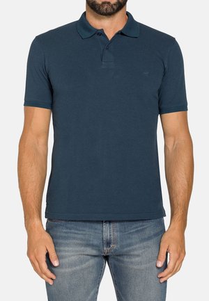 Uomo che indossa una polo blu scuro a maniche corte con colletto e due bottoni, abbinata a jeans azzurri.