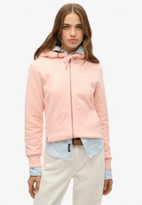 Sudadera de cremallera de color rosa claro con un bolsillo frontal, capucha ajustable con cordón y puños acanalados, superpuesta sobre una camisa de rayas azul.
