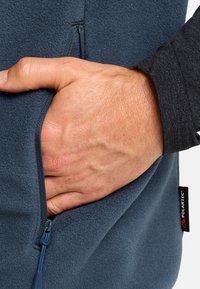 Fleecejacke in Dunkelblau mit einer Reißverschlusstasche. Nahaufnahme zeigt eine Hand, die in der Tasche ruht und die Textur sowie die Stoffdetails hervorhebt.