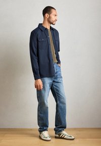 Man standing indoors wearing a navy button-up shirt jacket, olive green t-shirt, blue jeans, and beige and green sneakers.
