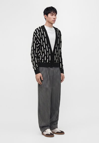 Homme portant un cardigan à motifs noir et blanc, une chemise blanche, un pantalon ample à fines rayures et des baskets blanches et marron, debout devant un fond uni.